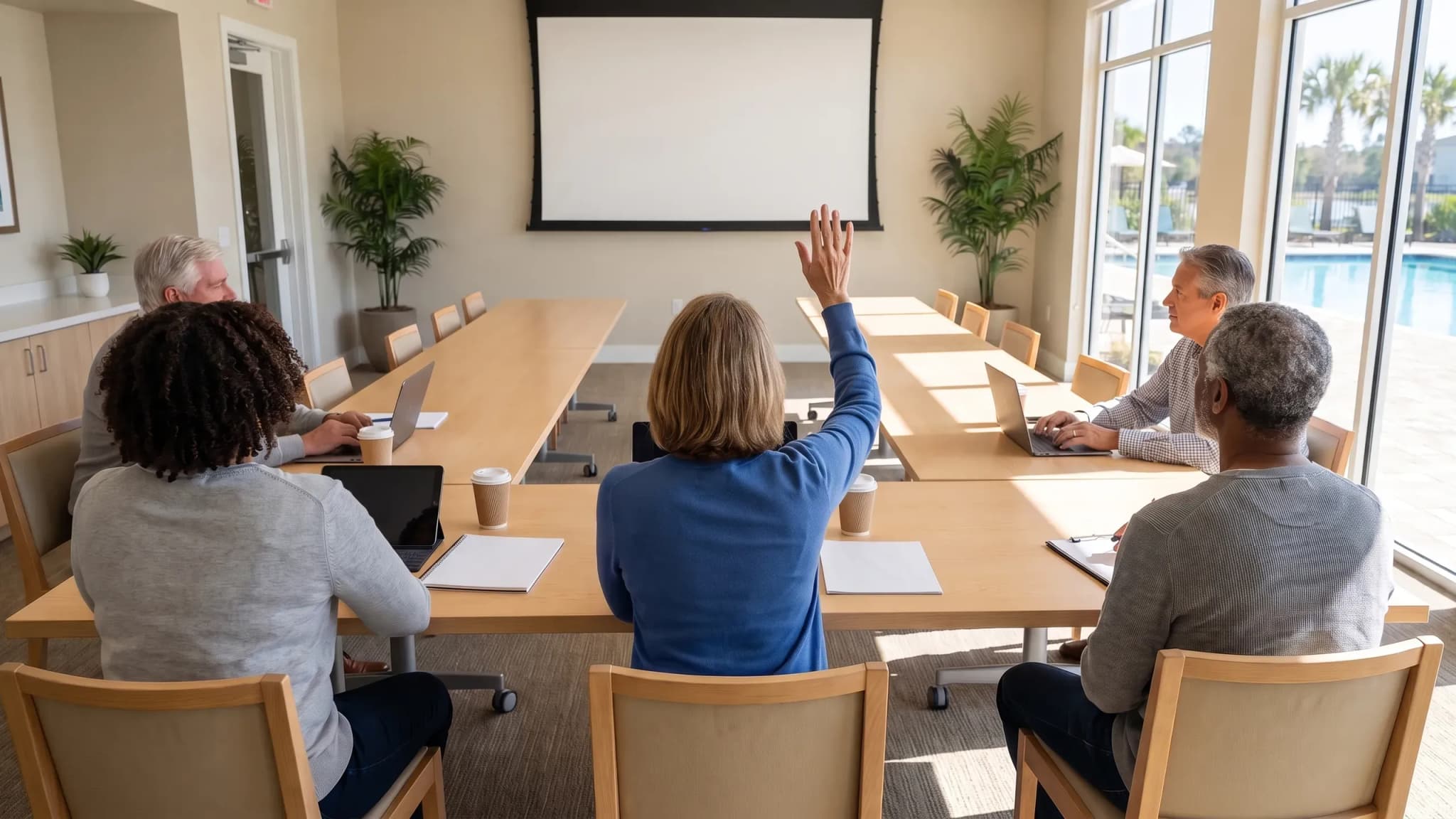 Five board members sit around one long table in a bright clubhouse meeting room, one raising a hand to vote, with a blank projector screen and a pool beyond the windows.