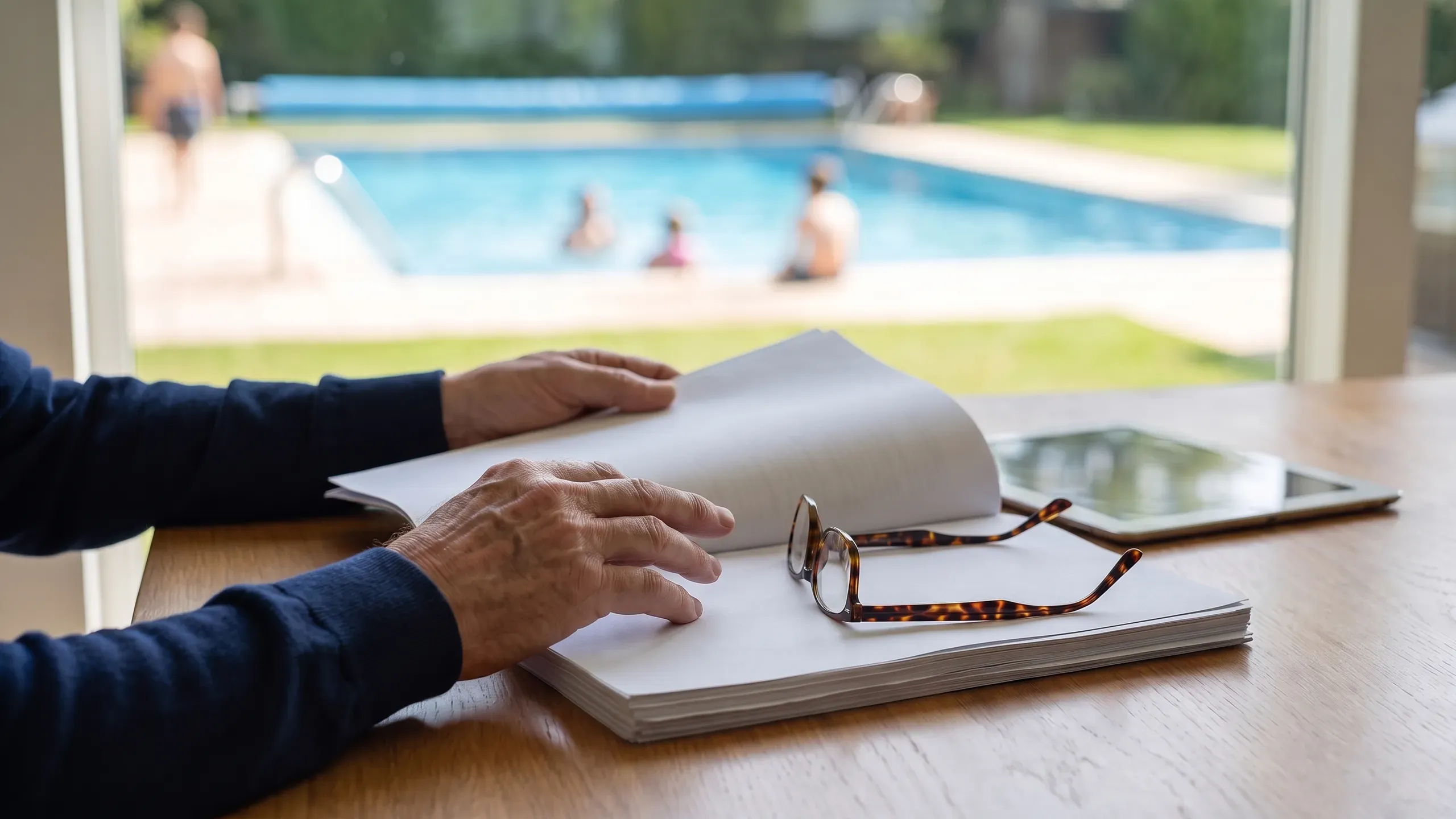 Hands of a board member rest on a printed proposal packet with folded reading glasses and a tablet beside it, a clear blue community pool soft through the window.