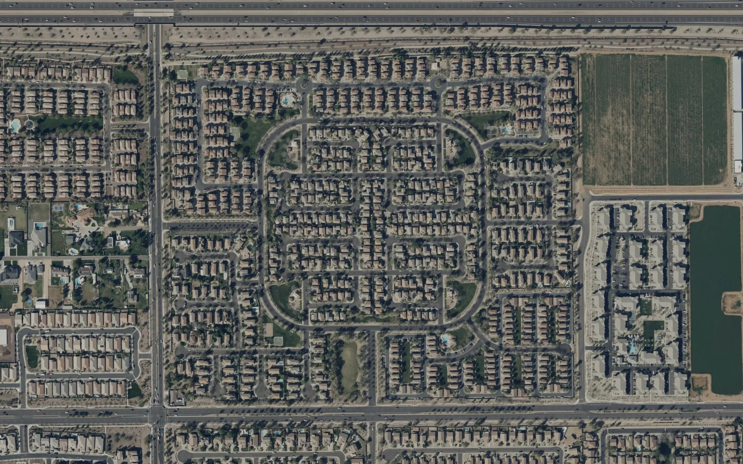 Satellite view of an Arizona community: tan-roofed single-family homes on curving desert blocks, with a pool and green commons inside it and a farm field beyond one edge.