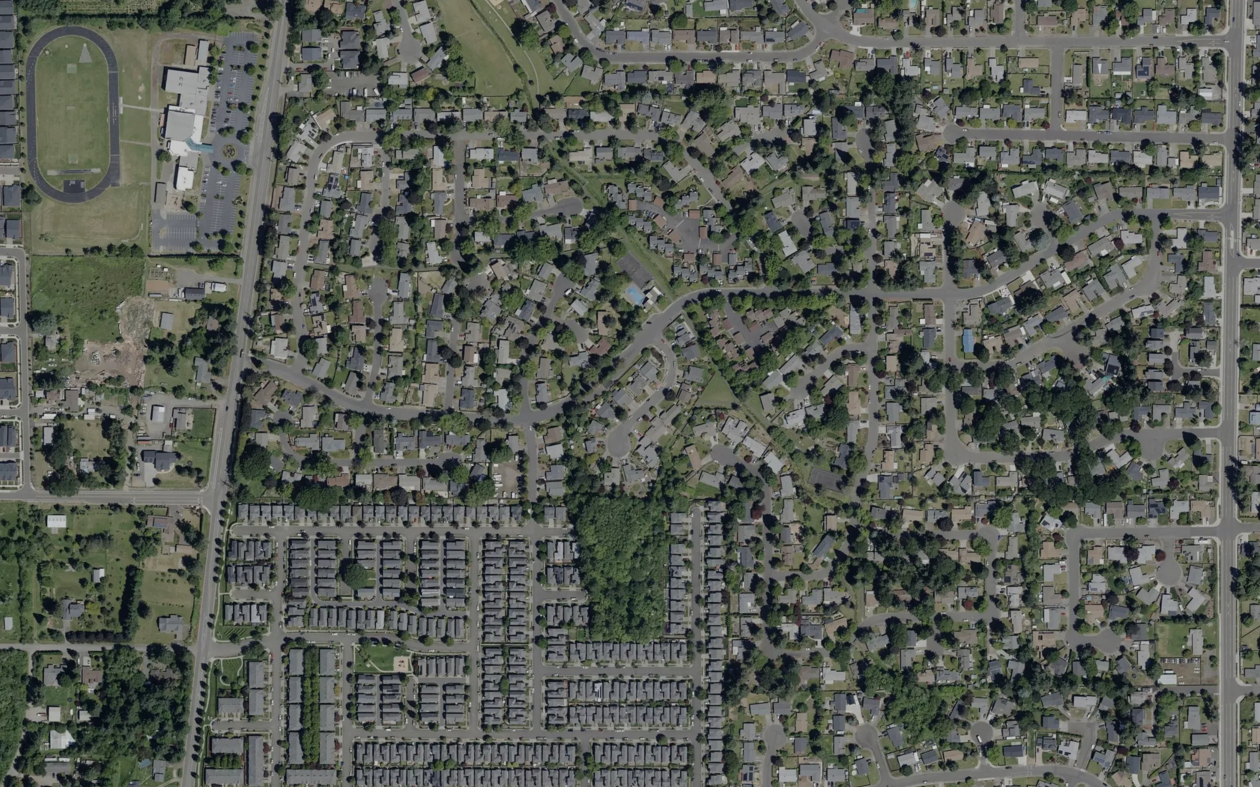 Satellite view of an Oregon community: detached houses on curving streets under heavy tree cover, with a school running track in one corner of the frame and rows of apartment blocks along the lower edge.