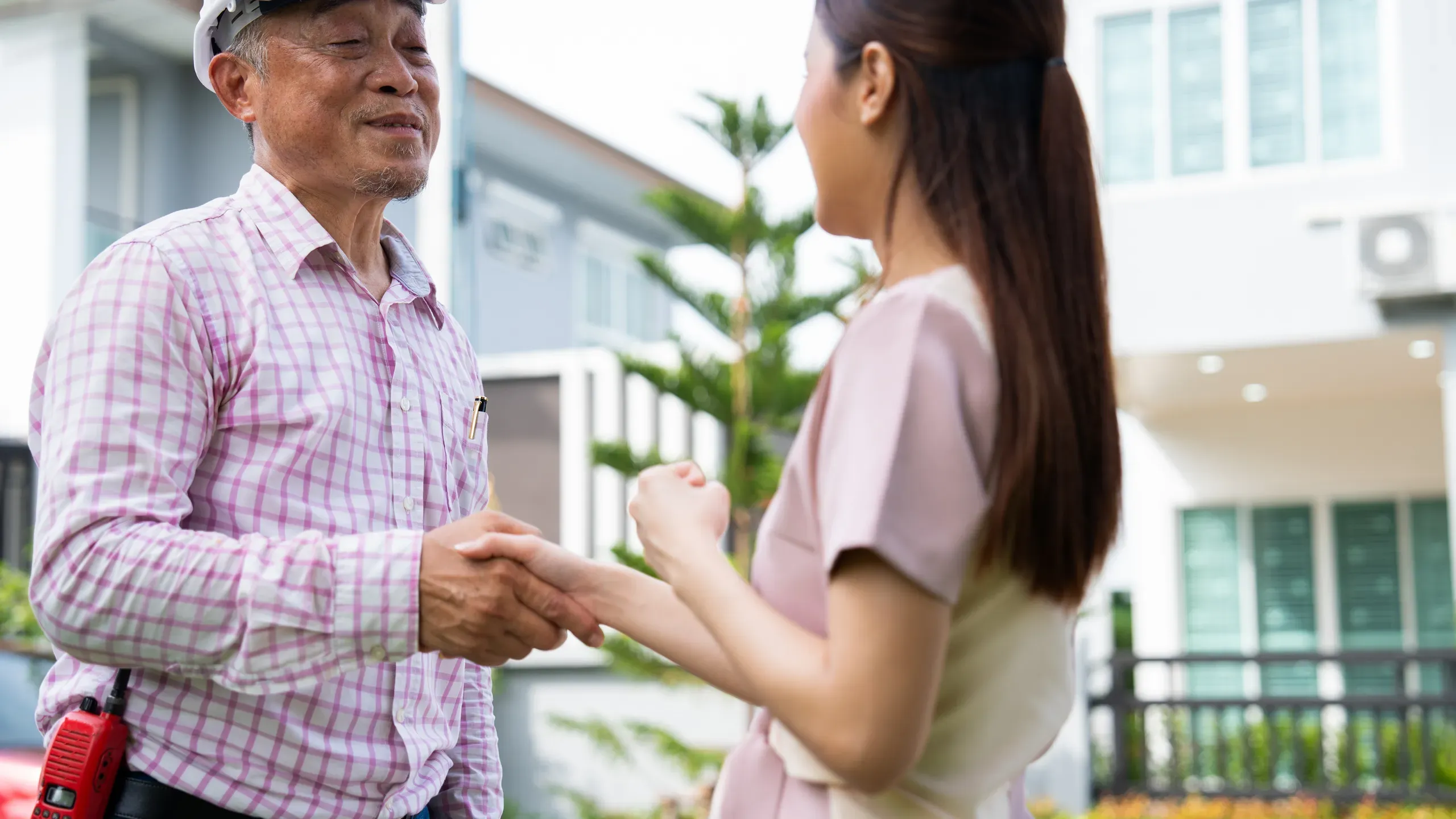 A smiling contractor in a pink checked shirt with a radio on his belt shakes hands with a homeowner in front of a white house.