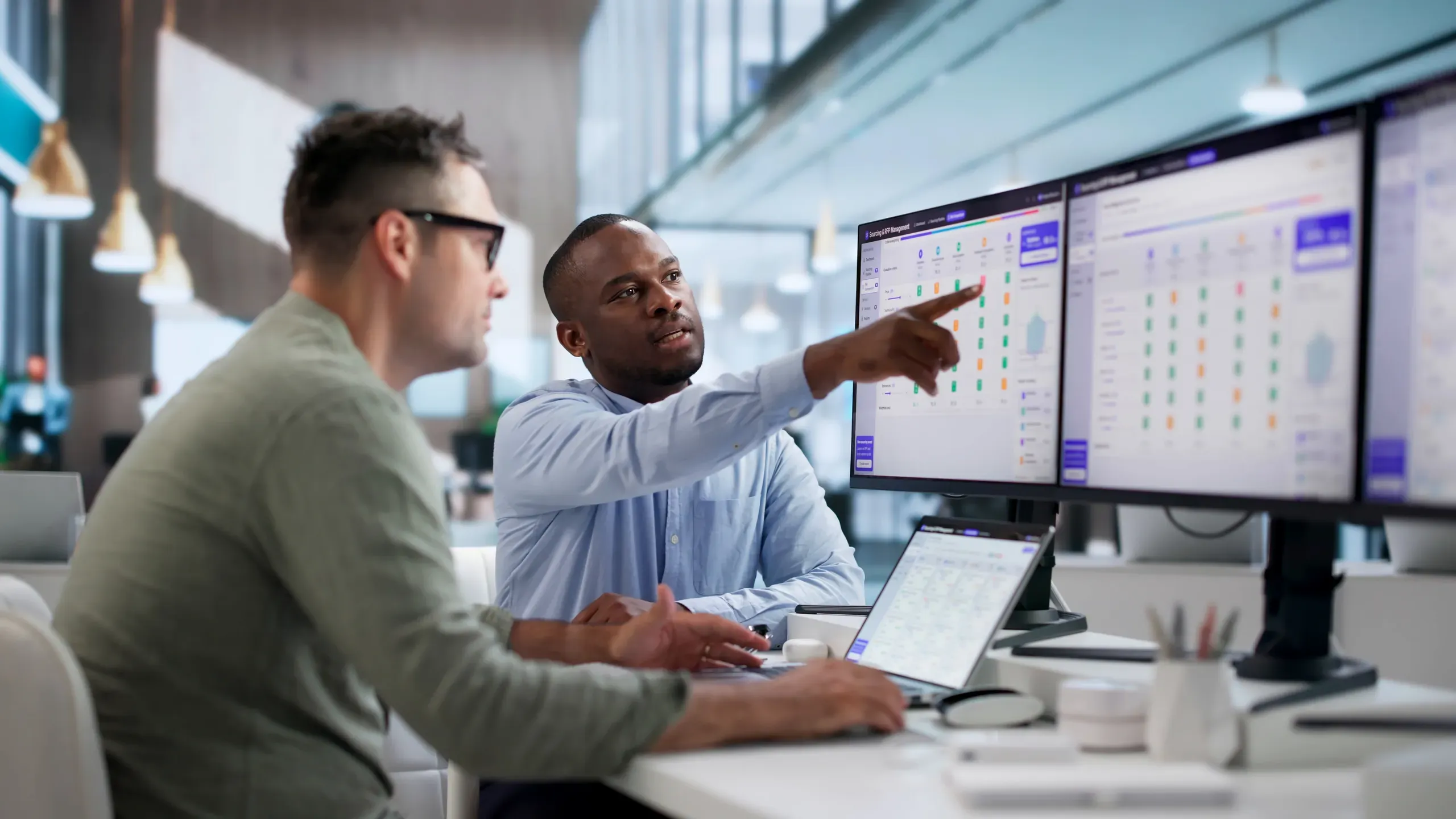 Two colleagues at a desk look at a wide monitor showing a grid of vendor proposals while one points at the screen, a laptop open in front of them.