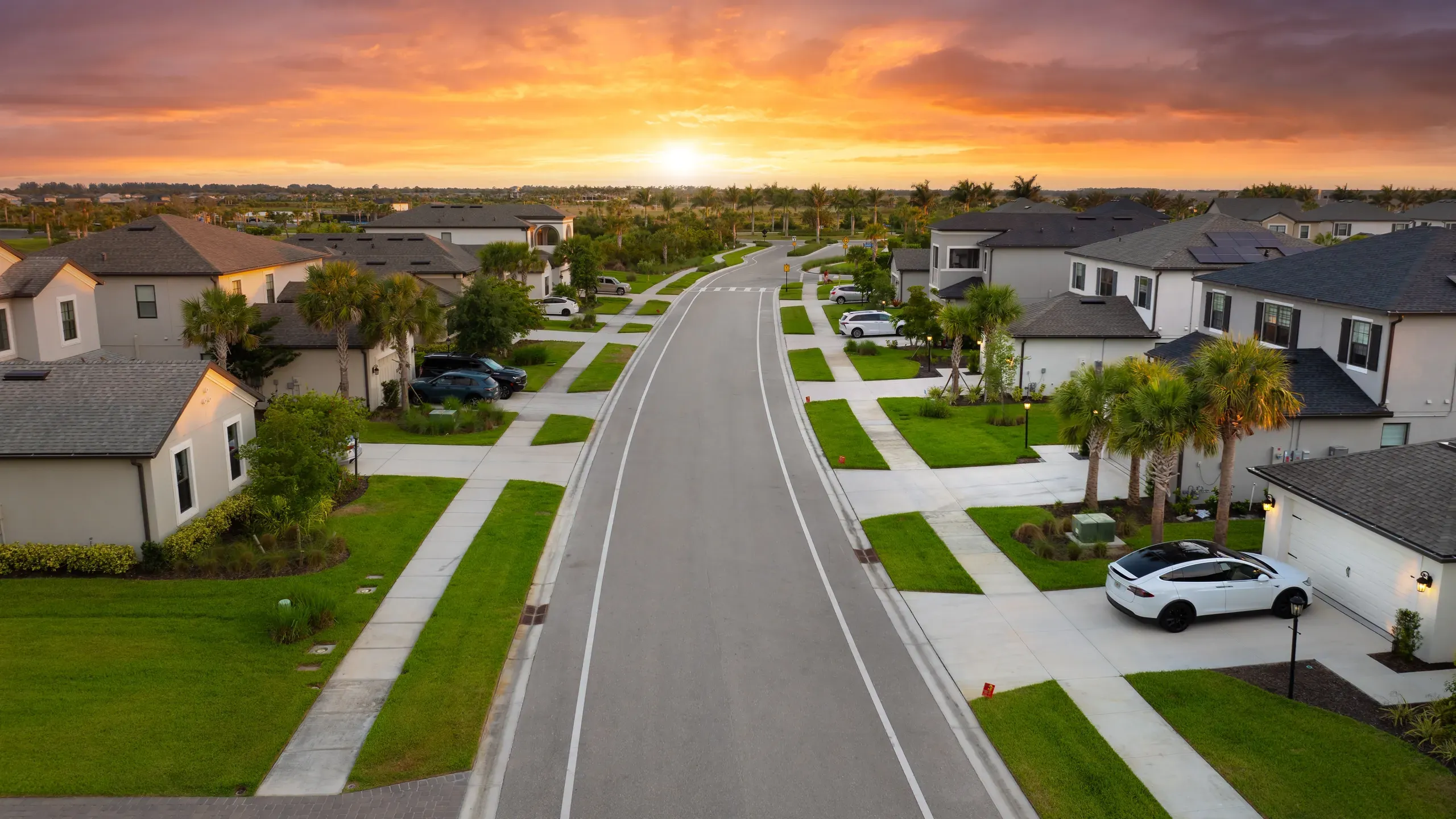 A low aerial view down a wide street lined with homes and palms in a southwest Florida neighbourhood under a vivid orange sunset sky.
